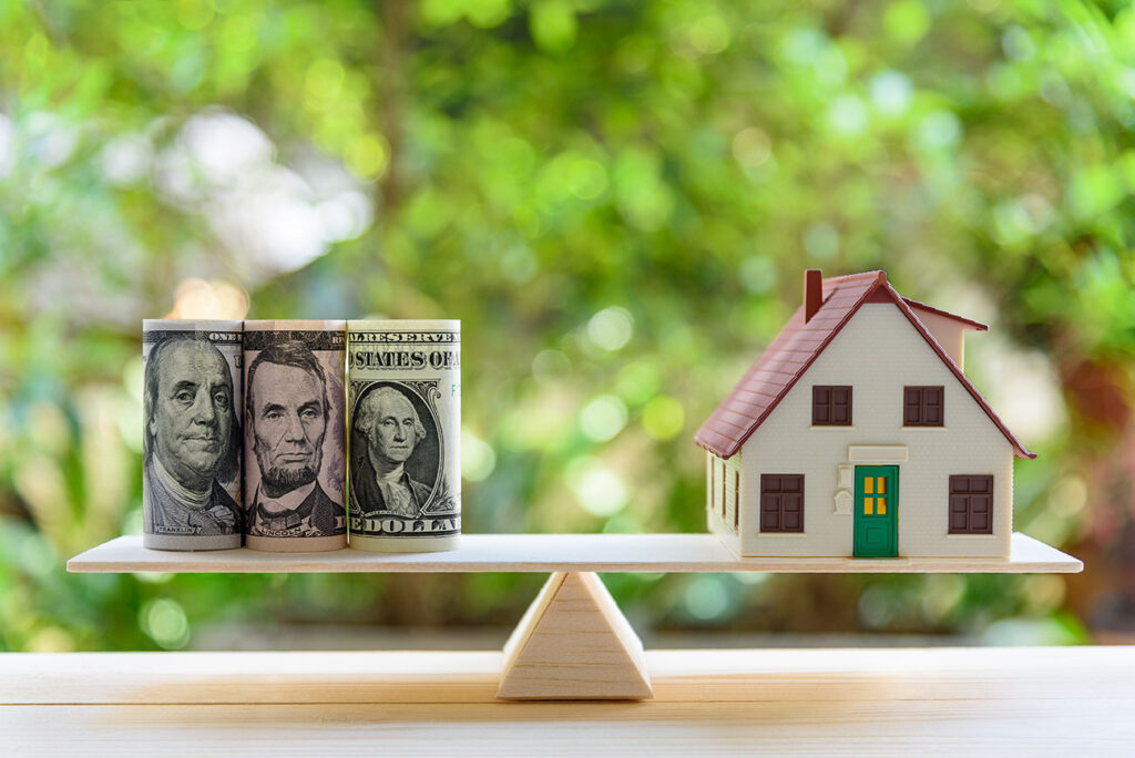 A wooden scale with money on the left being balanced by a small model house.