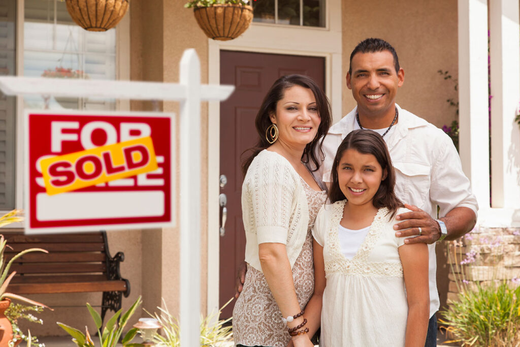 Family standing next to a For Sale sign. There is a sticker over the sign that says 'Sold'