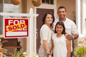A smiling family standing in front of their first home. There is a sign that says the house was just sold.