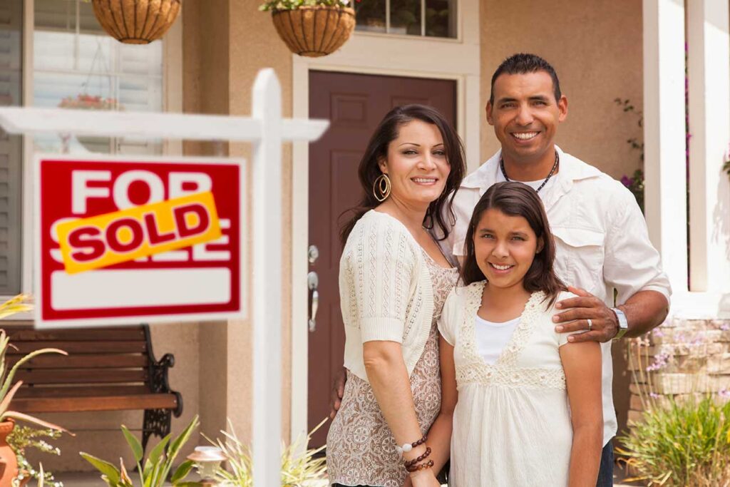 Family who just purchased their first house standing next to the 'For Sale' sign.